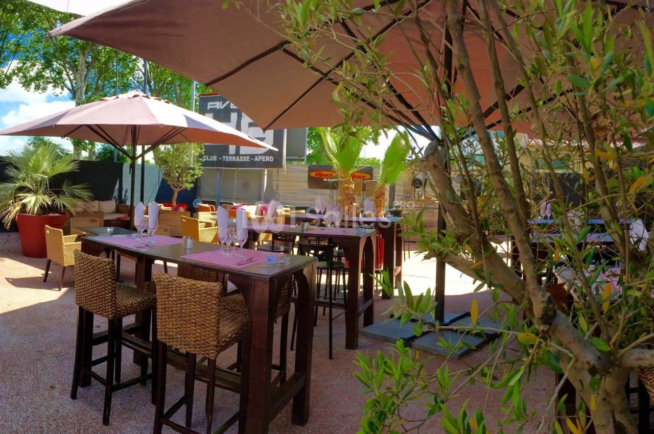 Terrasse extérieure avec tables hautes, chaises en osier et parasols, entourée de plantes et d'arbres.