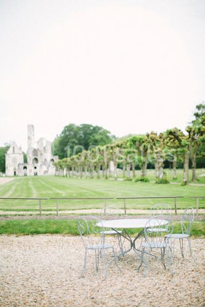 Table et chaises en fer forgé sur un sol gravillonné, avec une pelouse, des arbres taillés et des ruines en arrière-plan.