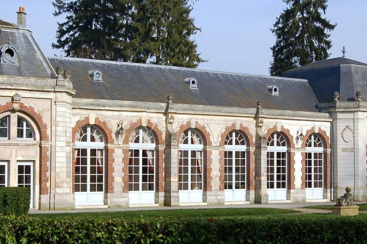 Façade d'un bâtiment ancien avec des arcades vitrées, entouré de végétation et de grands arbres.