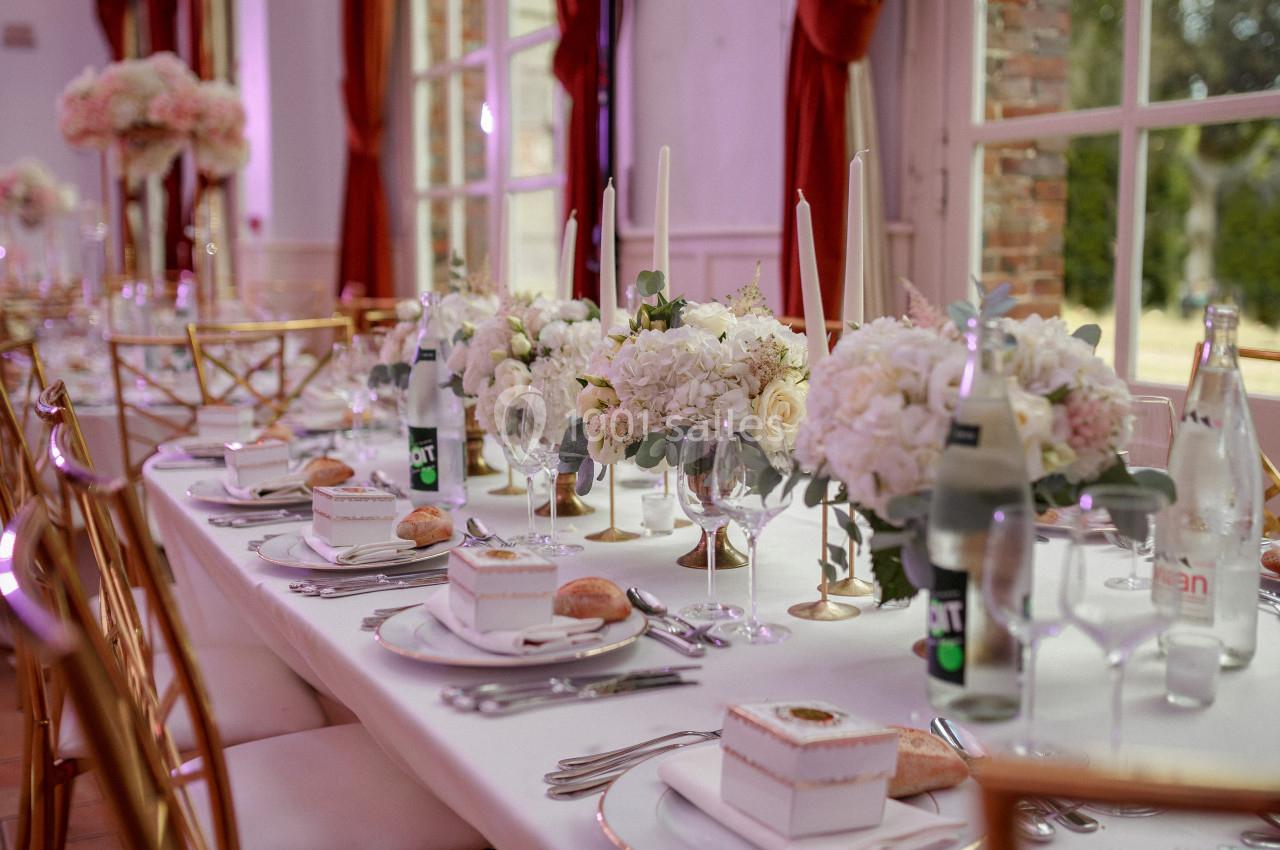 Table élégamment dressée avec nappes blanches, fleurs, chandeliers et vaisselle raffinée dans une salle lumineuse.