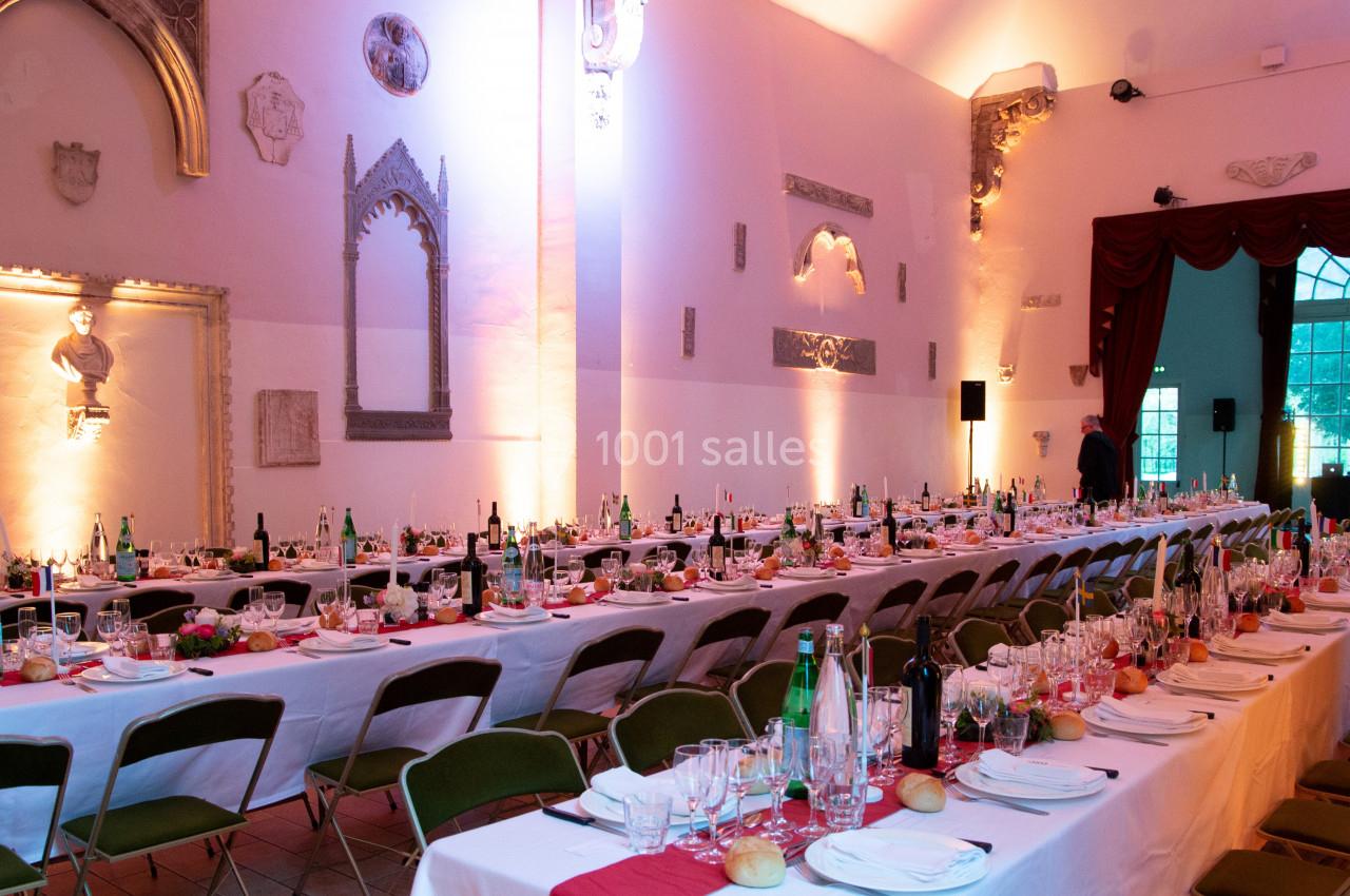 Salle de réception décorée avec de longues tables dressées, ornées de fleurs et de bouteilles, éclairage chaleureux.