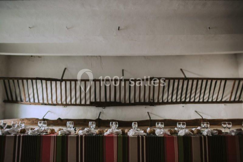 Table décorée avec des verres, serviettes et pains, placée devant un mur orné d'outils agricoles anciens.