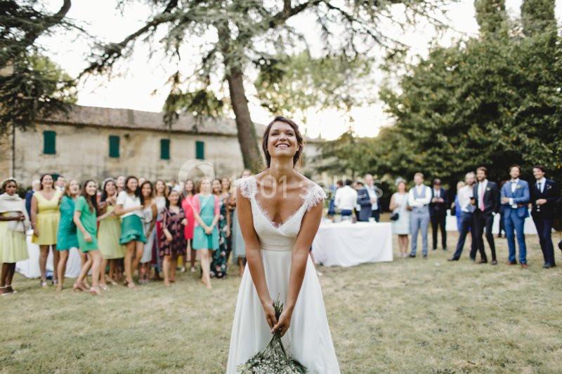 Une mariée souriante s'apprête à lancer son bouquet devant un groupe d'invités dans un jardin.
