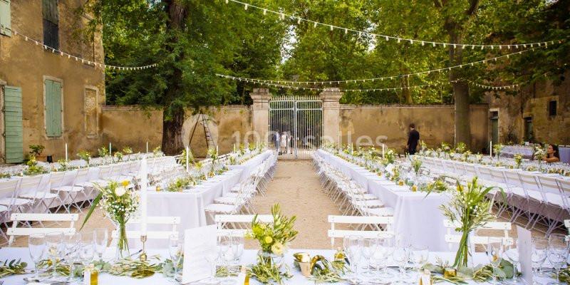 Tables décorées pour un mariage en extérieur, disposées en rangées sous des guirlandes lumineuses dans une cour arborée.