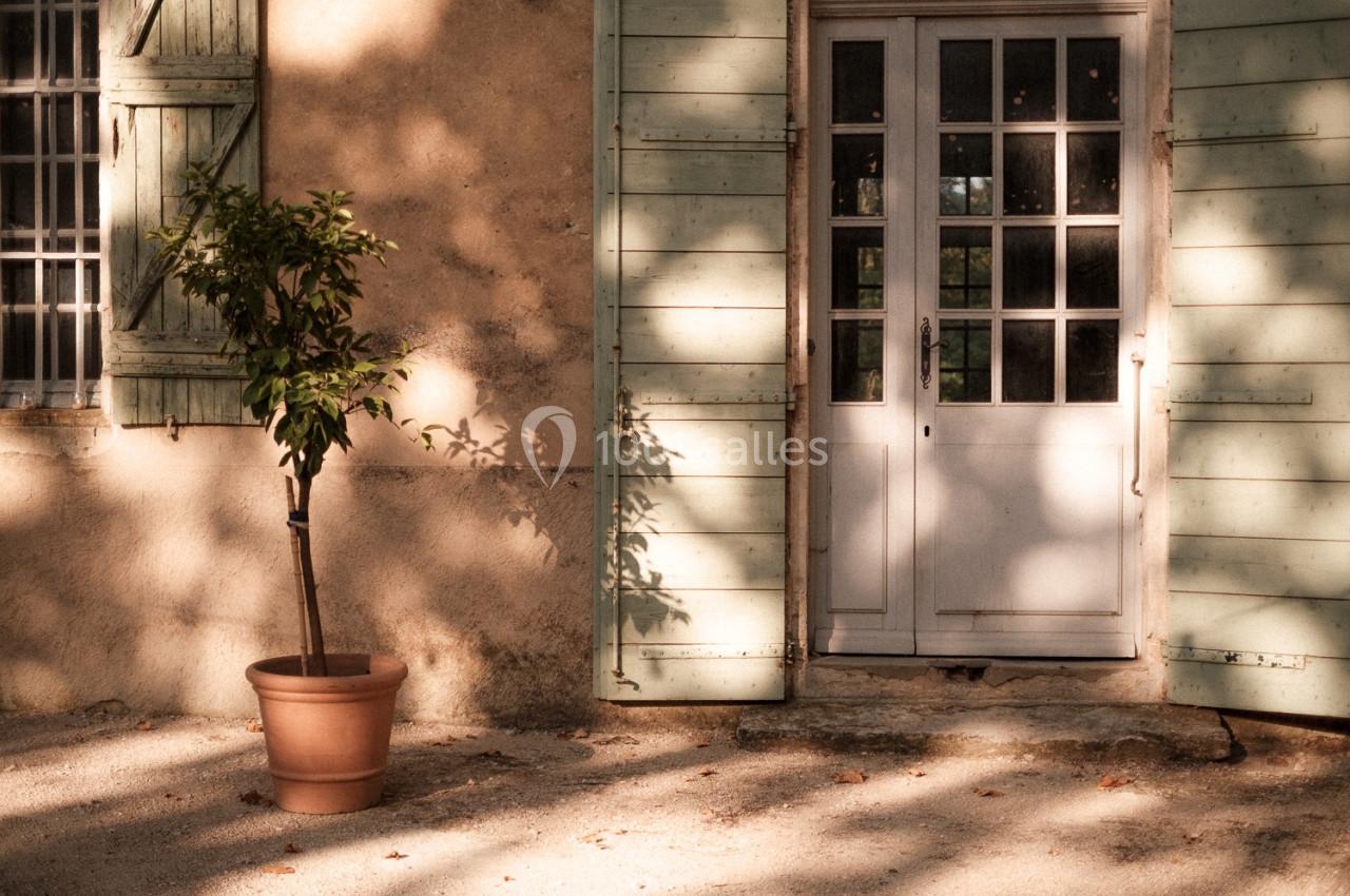 Façade ensoleillée d'une maison avec volets verts, une porte vitrée et un petit arbre en pot sur le sol.
