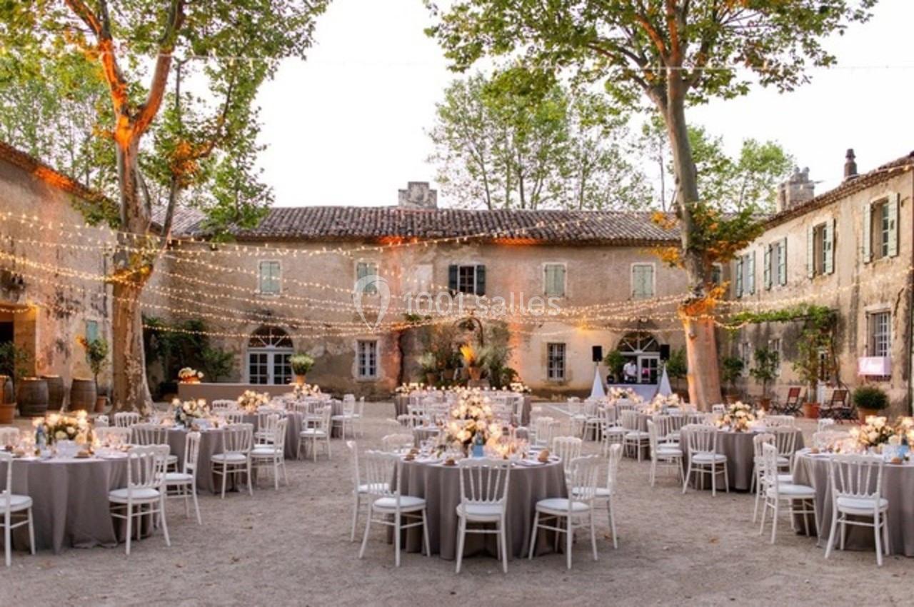 Cour d'un domaine ancien décorée pour un mariage, avec tables dressées, chaises blanches et guirlandes lumineuses suspendues.