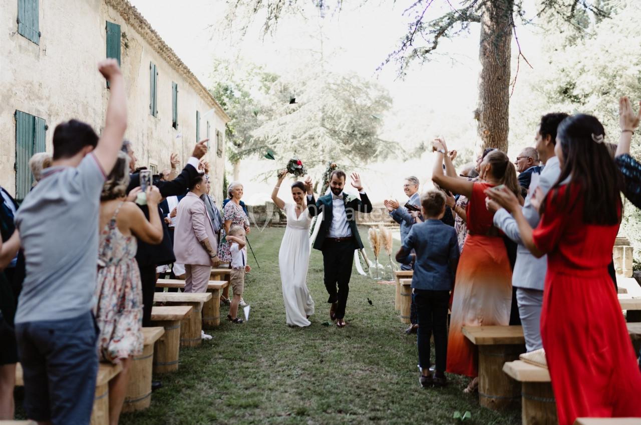 Un couple de mariés marche joyeusement entre des invités applaudissant dans un jardin lors d'une cérémonie en plein air.