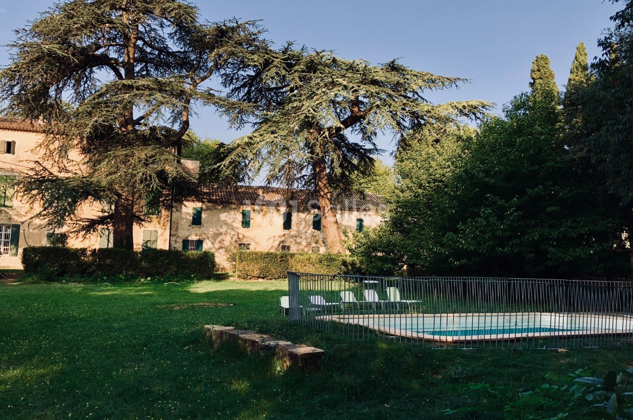 Jardin verdoyant avec piscine clôturée, grands arbres et bâtiment ancien en arrière-plan sous un ciel dégagé.