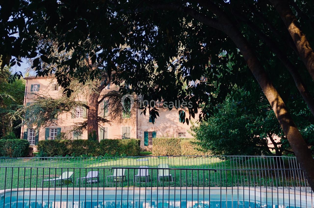 Maison en pierre entourée de verdure avec une piscine clôturée au premier plan, vue à travers des arbres.