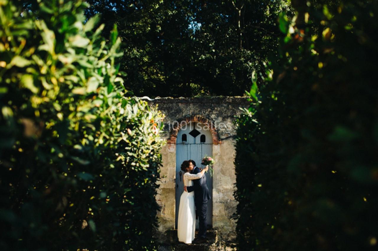 Un couple en tenue de mariage s'embrasse devant une porte ancienne, entouré de haies verdoyantes.