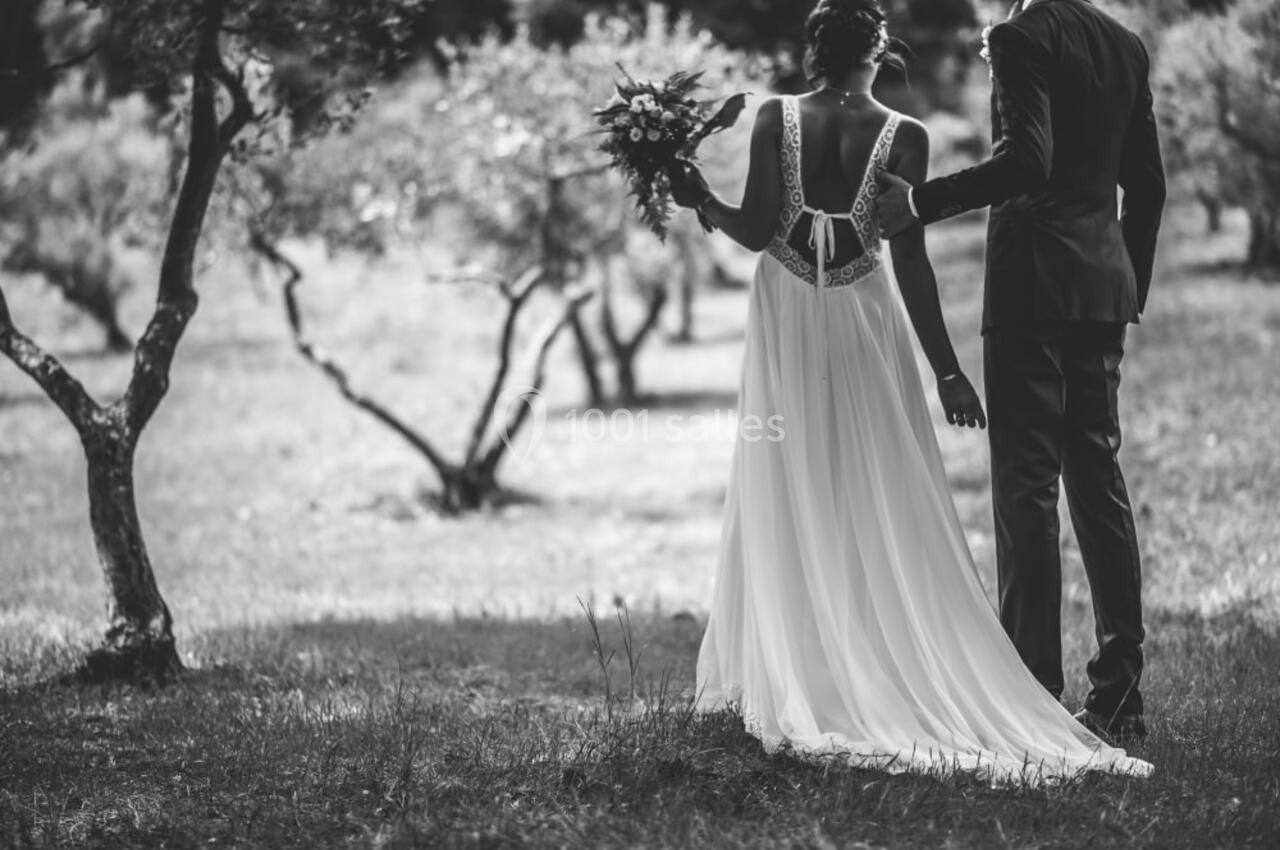 Un couple en tenue de mariage se tient debout dans un jardin, entouré d'arbres, vu de dos.