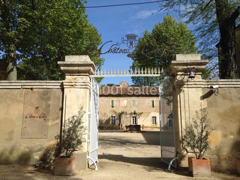 Entrée d'un domaine avec portail blanc ouvert, murs en pierre et inscription ’Château Clos’ sous un ciel bleu.