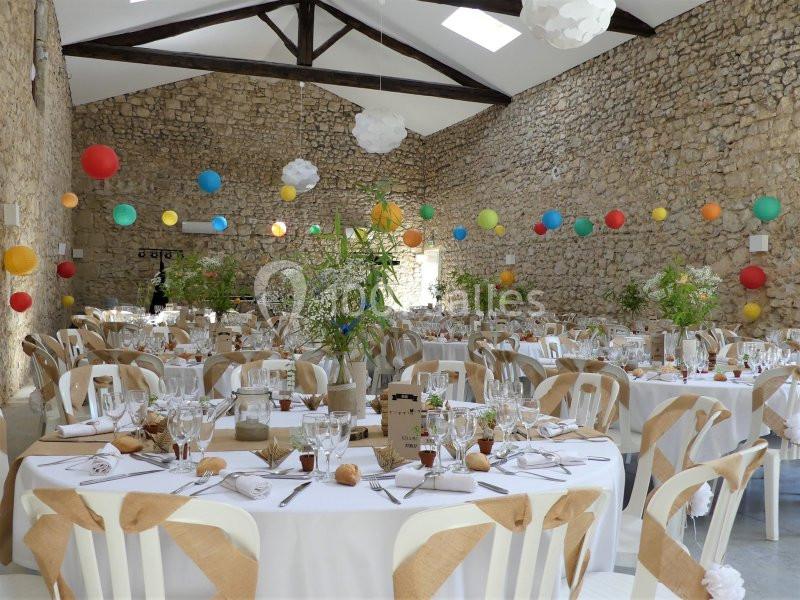 Salle de réception décorée avec des guirlandes colorées, des tables rondes dressées et des murs en pierre apparente.