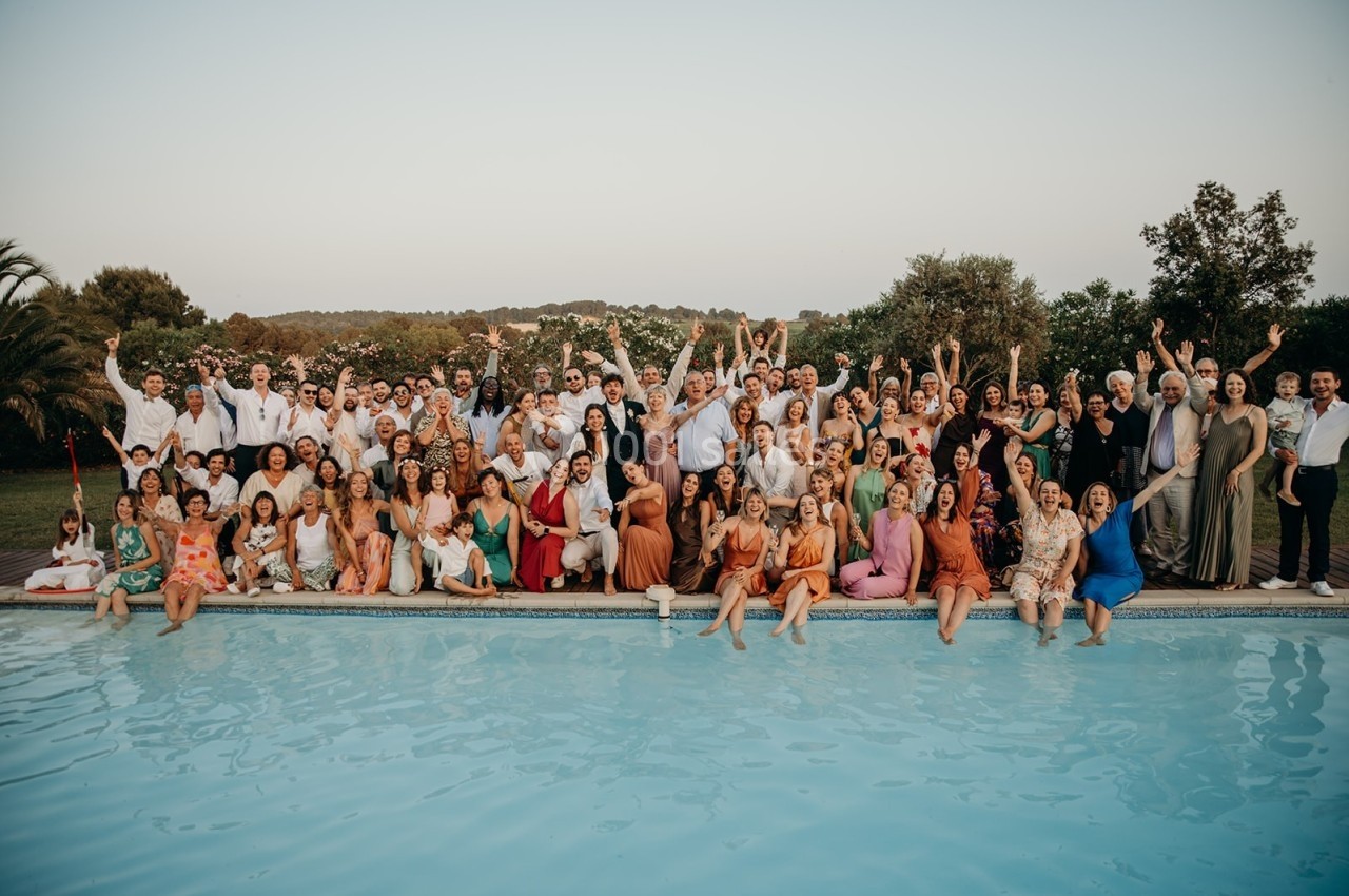 Un grand groupe de personnes posant joyeusement au bord d'une piscine, entouré de végétation et d'un ciel dégagé.