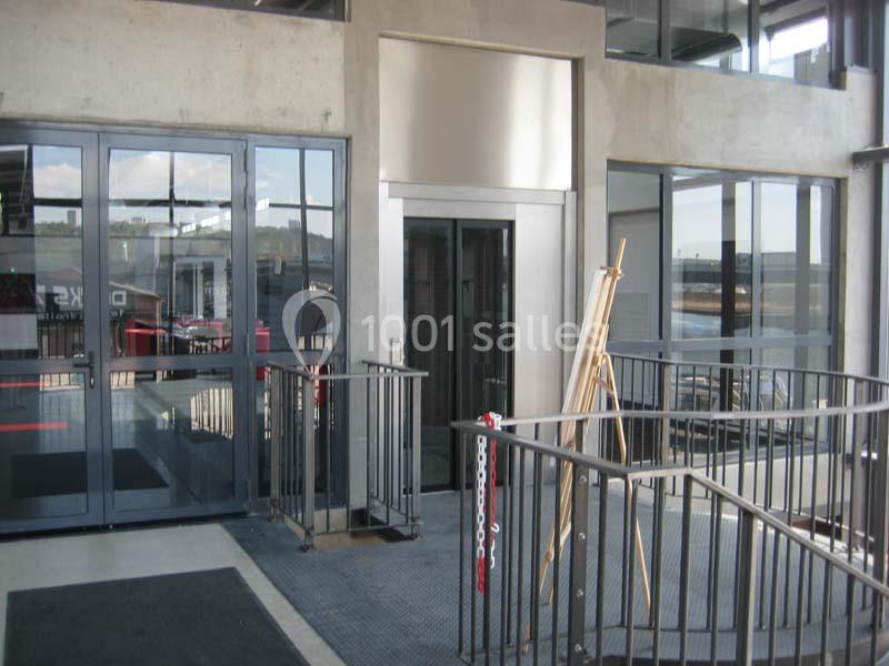 Entrée d'un bâtiment avec une porte vitrée, un ascenseur et des rampes d'accès en métal.