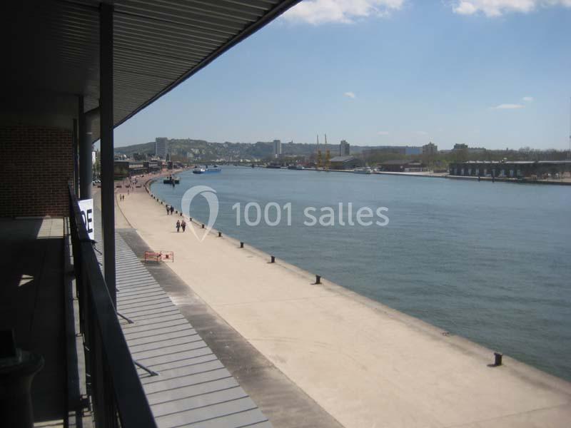 Vue d'un quai longeant une rivière sous un ciel dégagé, avec des bâtiments et collines en arrière-plan.