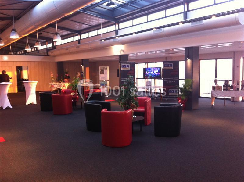 Espace intérieur lumineux avec fauteuils rouges et noirs, tables hautes, plantes et stands d'exposition en arrière-plan.