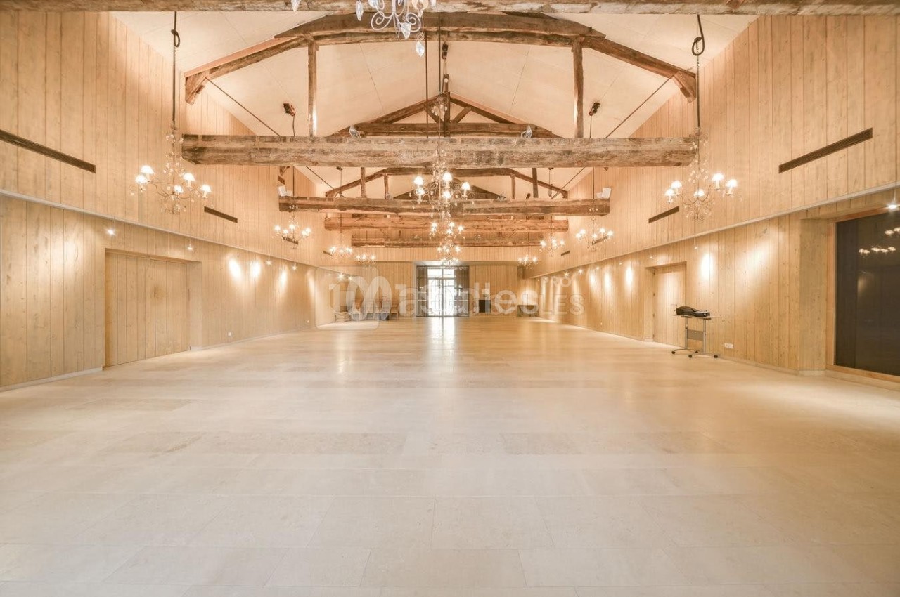 Grande salle lumineuse avec poutres apparentes, murs en bois clair, lustres suspendus et sol en pierre beige.