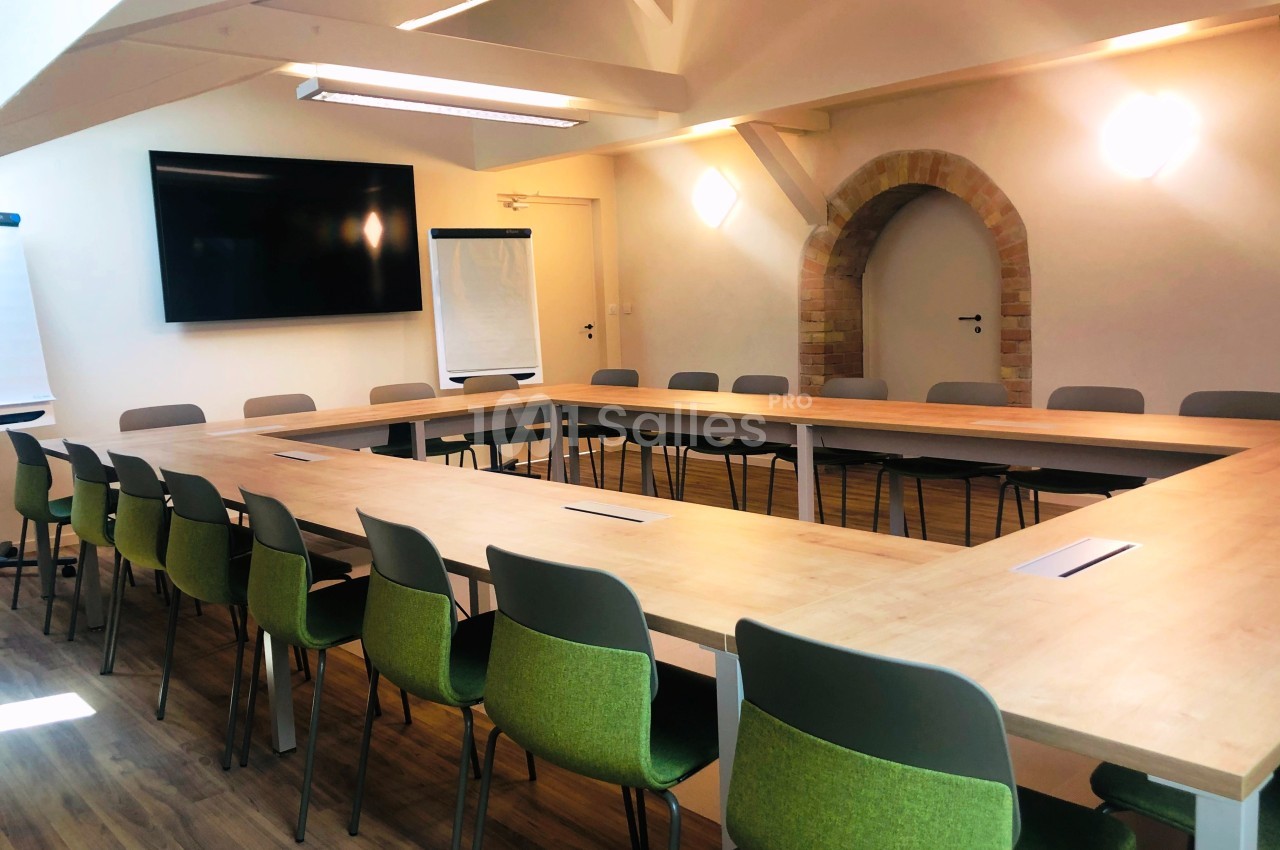 Salle de réunion lumineuse avec tables disposées en U, chaises vertes, écran mural et paperboard.