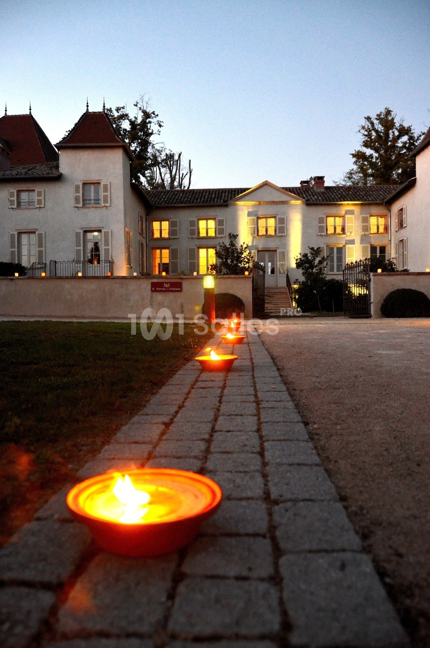 Allée pavée bordée de bougies allumées menant à une grande maison éclairée au crépuscule.