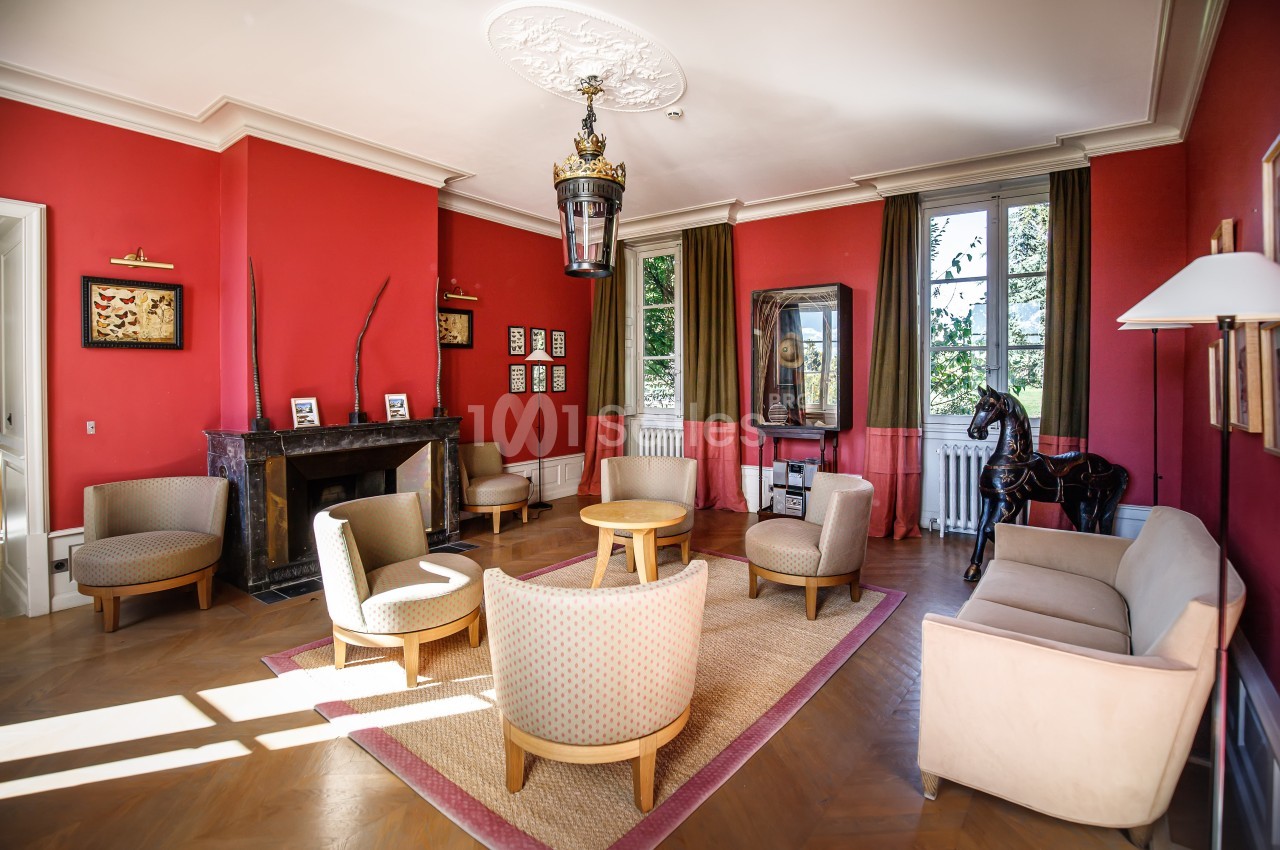 Salon lumineux avec murs rouges, cheminée en marbre, fauteuils disposés autour d'une table et grande fenêtre donnant sur un…