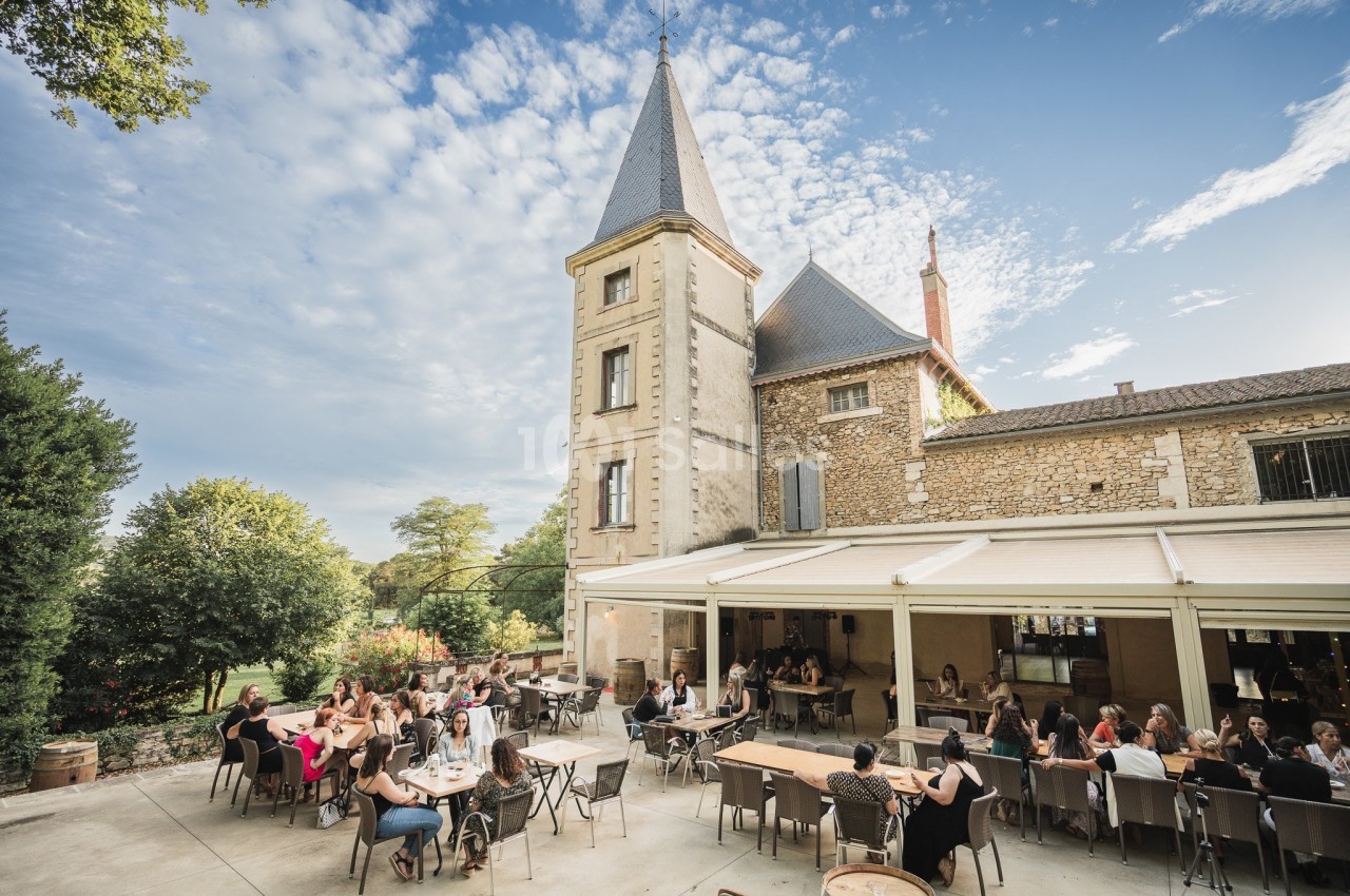 Groupe de personnes attablées en terrasse devant un bâtiment en pierre avec une tour, sous un ciel partiellement nuageux.