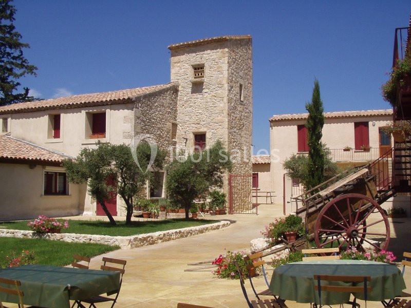 Cour intérieure d'un mas provençal avec une tour en pierre, des oliviers, des fleurs et une charrette en bois.