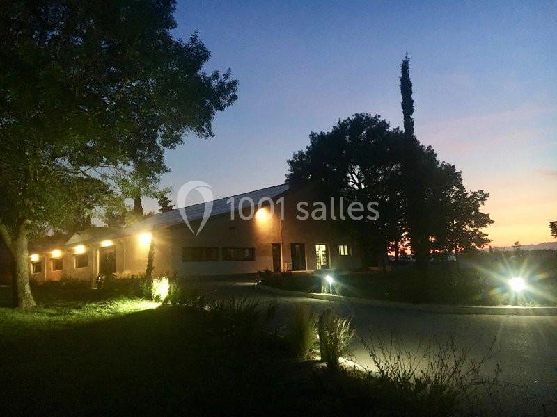 Bâtiment éclairé à la tombée de la nuit, entouré d'arbres et d'un chemin bordé de lumières.