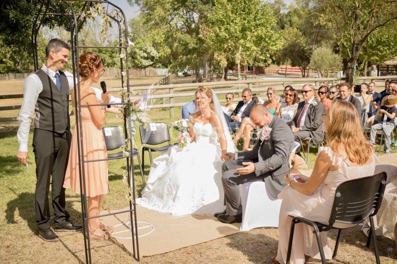 Cérémonie de mariage en plein air avec des invités assis, une mariée souriante et un officiant s'exprimant.