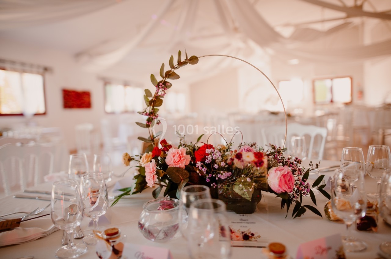 Centre de table floral avec roses et feuillages, entouré de verres et décorations dans une salle lumineuse.