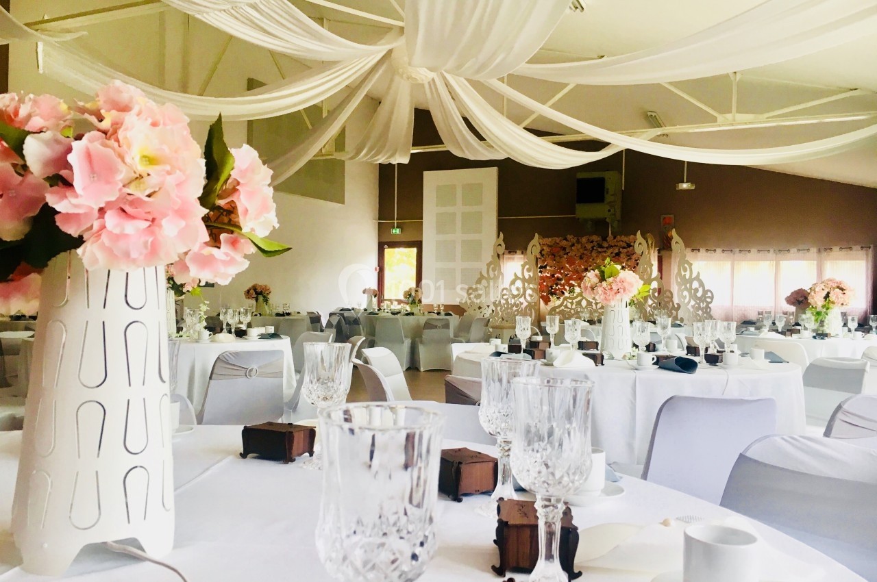 Salle de réception décorée avec des nappes blanches, des fleurs roses et des drapés suspendus au plafond.