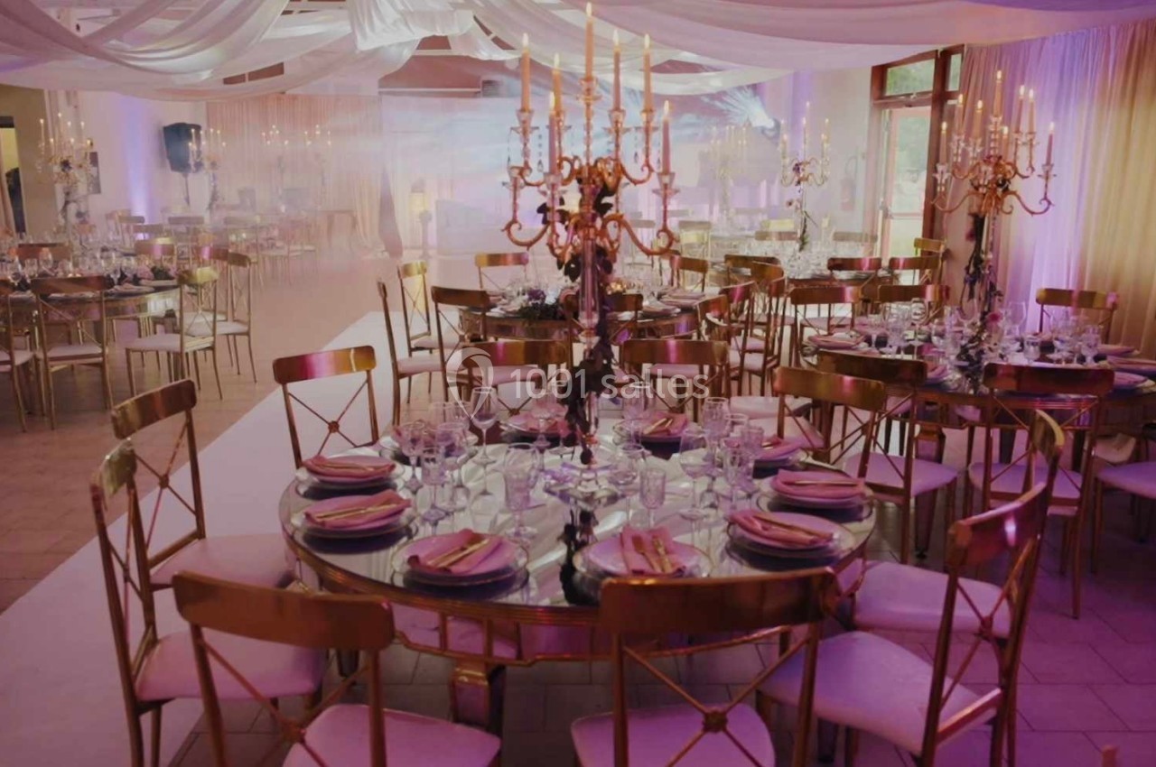 Salle de réception décorée avec des tables rondes, nappes élégantes, chandeliers dorés et éclairage tamisé.