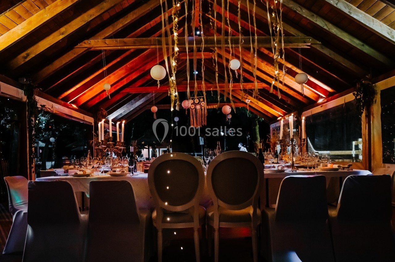 Salle décorée pour un événement, avec une table dressée, des chaises blanches et des lumières suspendues sous un toit en…