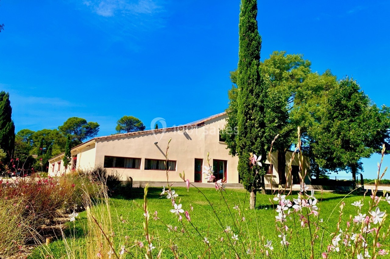 Bâtiment moderne entouré de pelouse, fleurs et arbres, sous un ciel bleu dégagé.