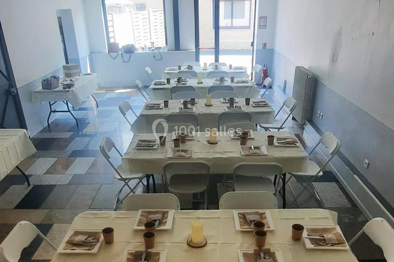 Salle aménagée avec plusieurs tables rectangulaires dressées, chaises blanches et lumière naturelle provenant des fenêtres.