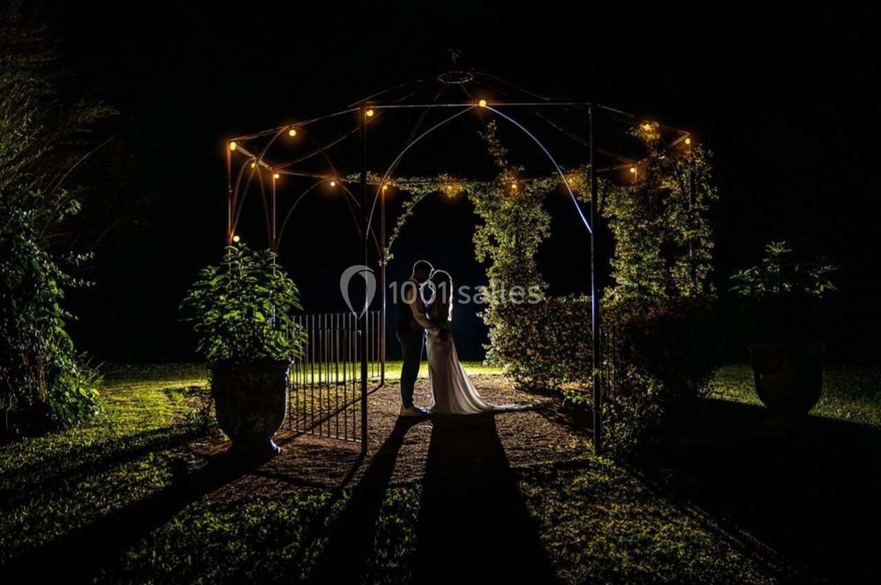 Un couple s'embrasse sous une arche illuminée par des guirlandes lumineuses dans un jardin nocturne.