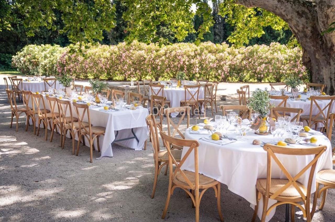 Tables dressées avec nappes blanches et décorations simples, disposées en extérieur sous des arbres et près de buissons…