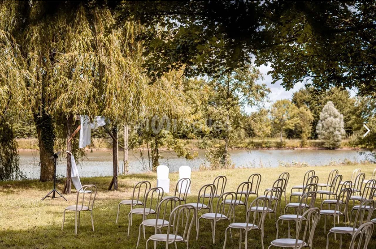 Chaises blanches disposées en extérieur sous des arbres, face à une rivière, pour un événement en plein air.