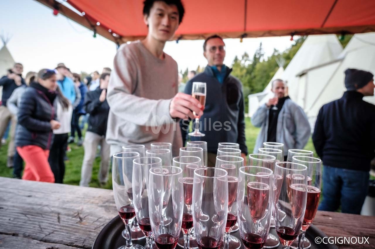 Un homme tend un verre de vin pétillant devant une table garnie de flûtes remplies, en extérieur sous une tente.