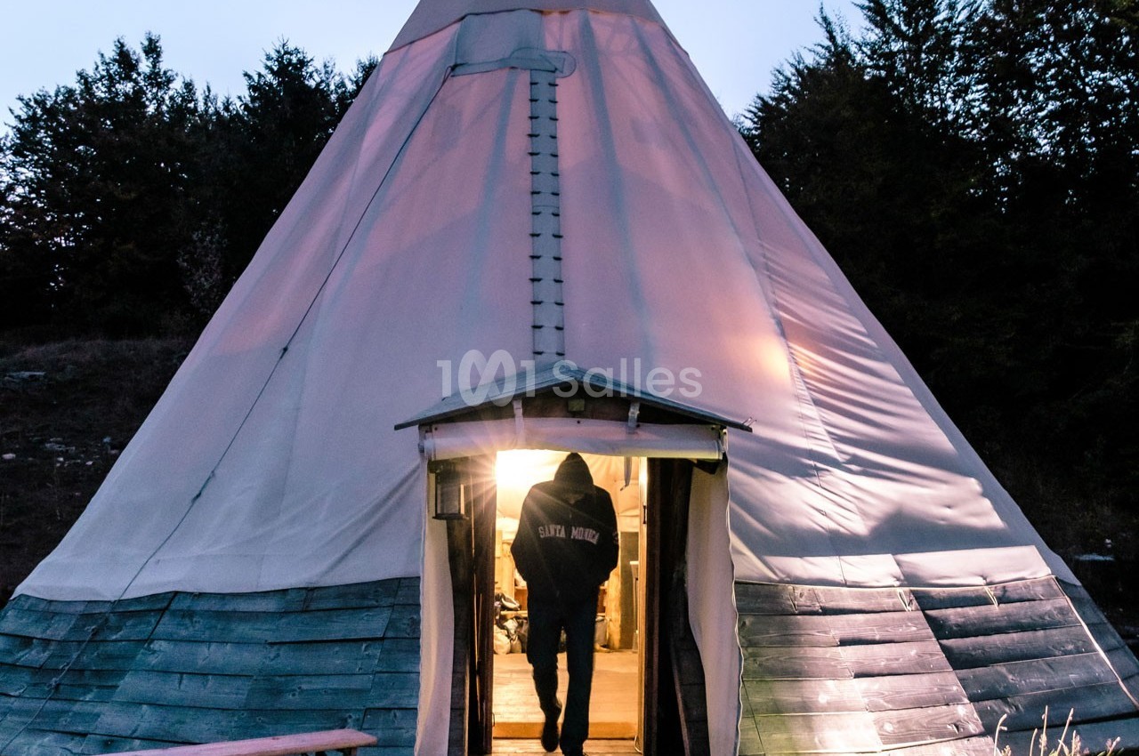 Un homme entre dans un tipi éclairé de l'intérieur, entouré d'arbres au crépuscule.