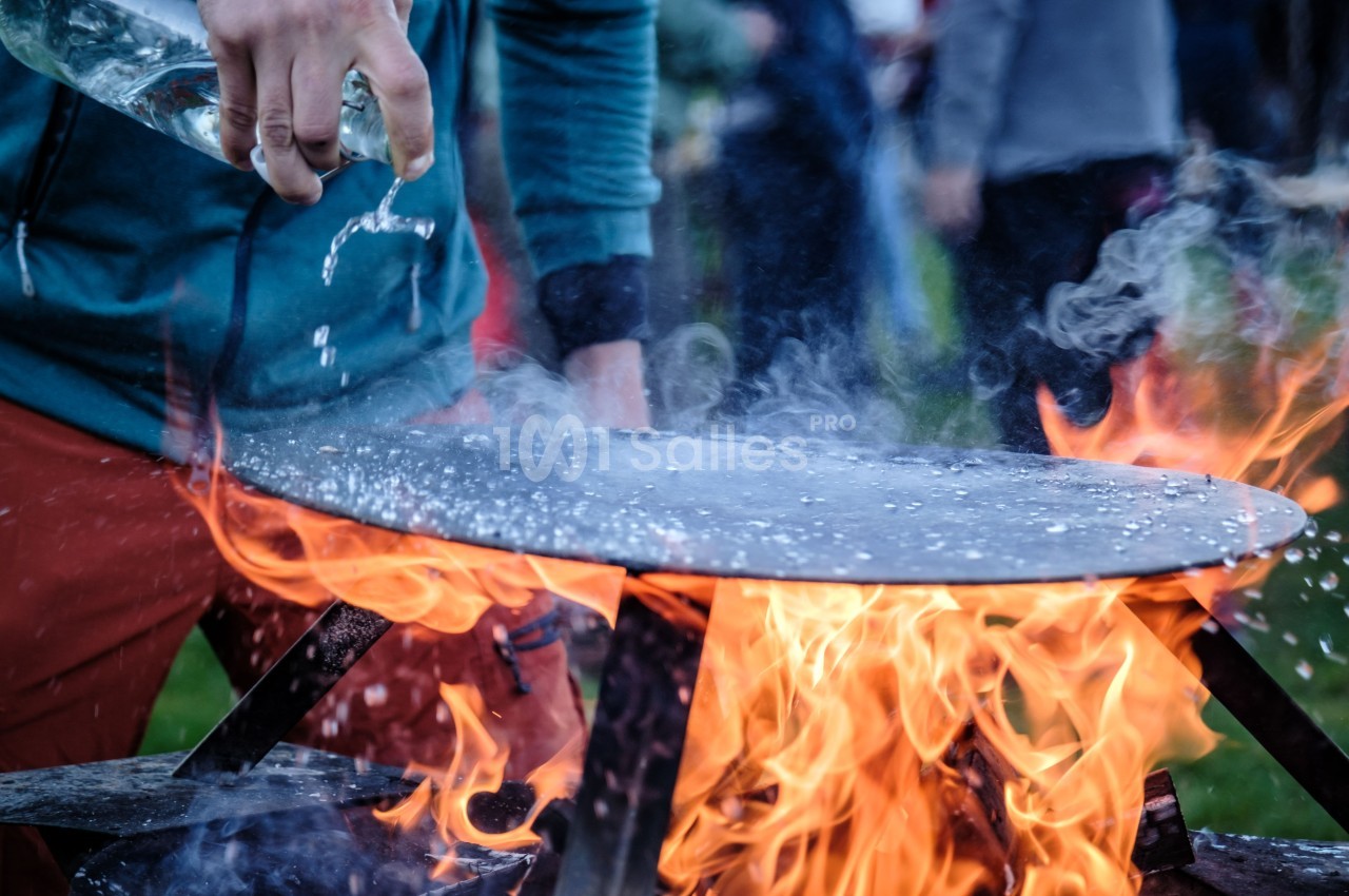 Une personne verse de l'eau sur une plaque chauffante au-dessus d'un feu vif en extérieur.