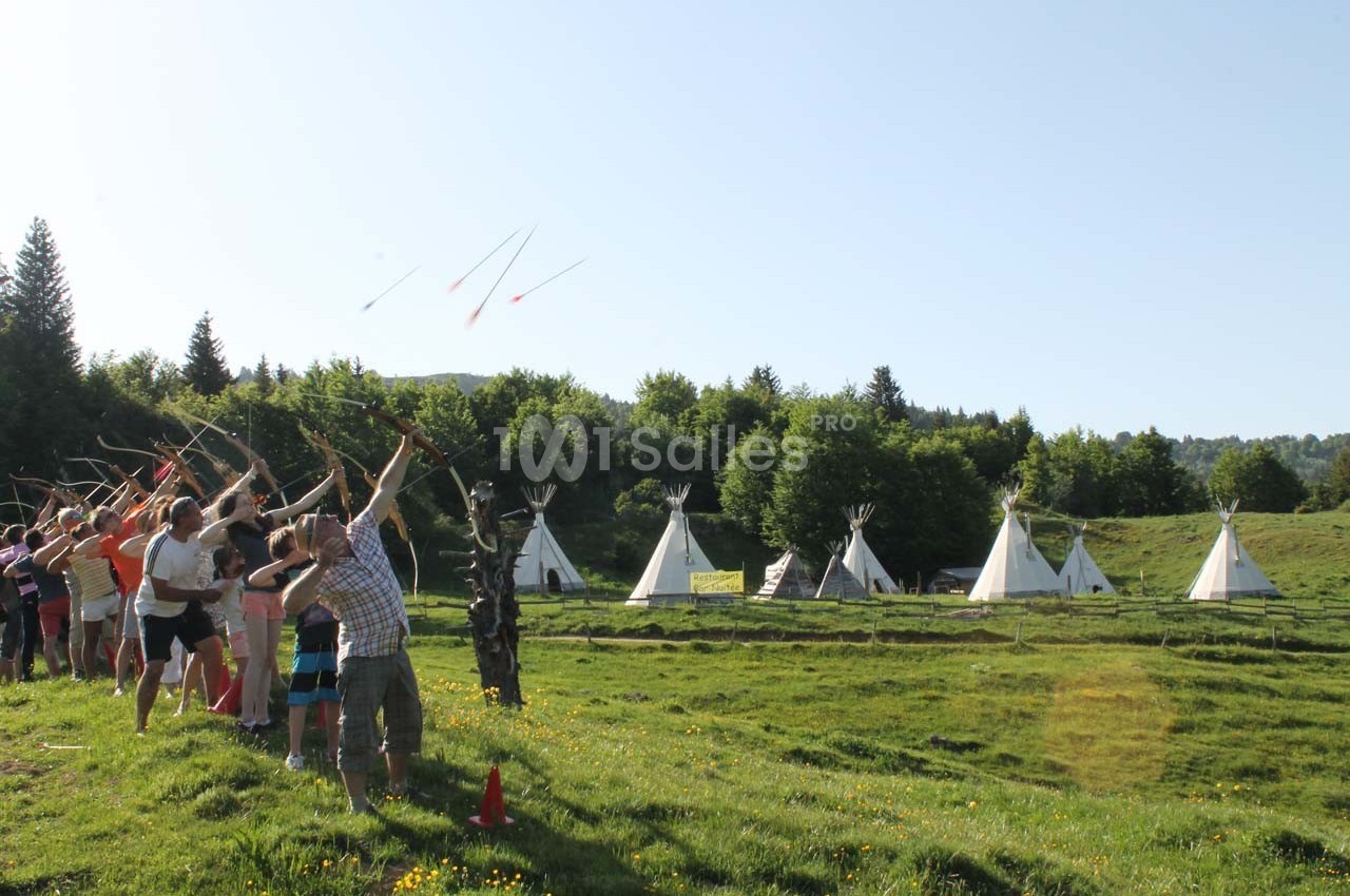 Un groupe de personnes lance des flèches avec des arcs dans un champ, près de tipis entourés de verdure.
