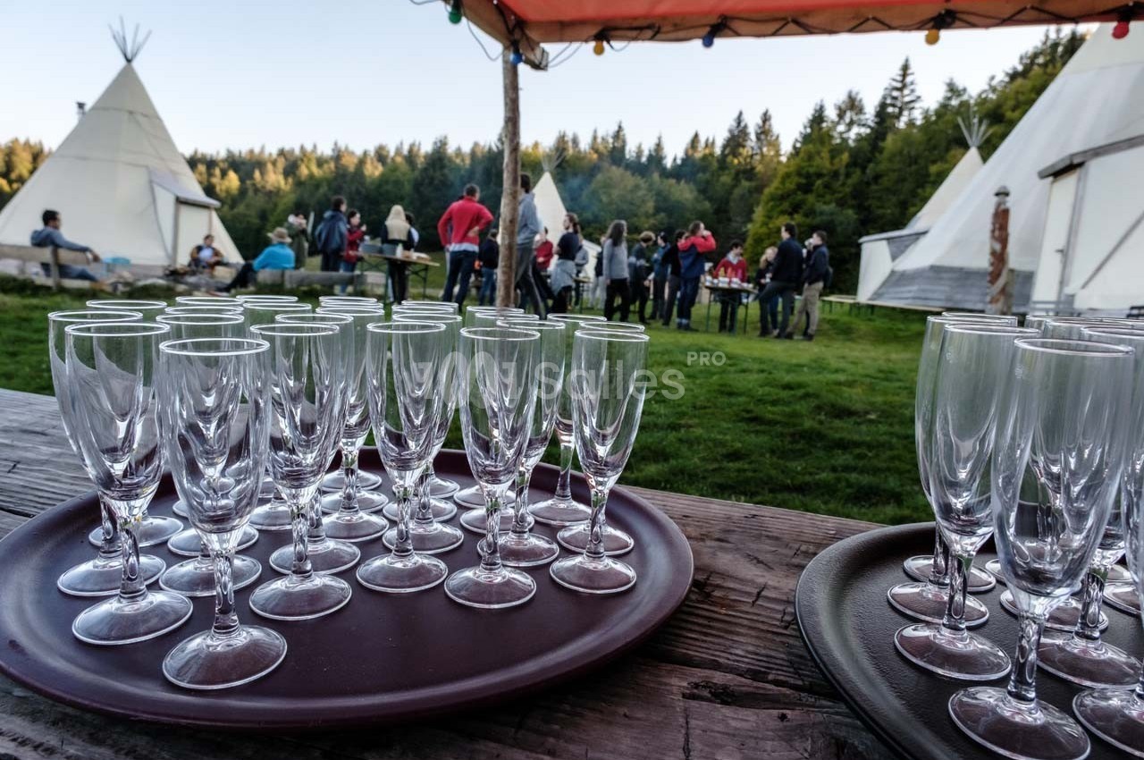 Plateaux de verres à pied posés sur une table en bois, avec des tipis et des personnes en arrière-plan dans un cadre naturel.