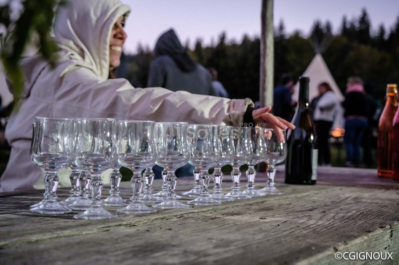 Une personne en veste claire dispose des verres sur une table en bois, avec un paysage extérieur en arrière-plan.