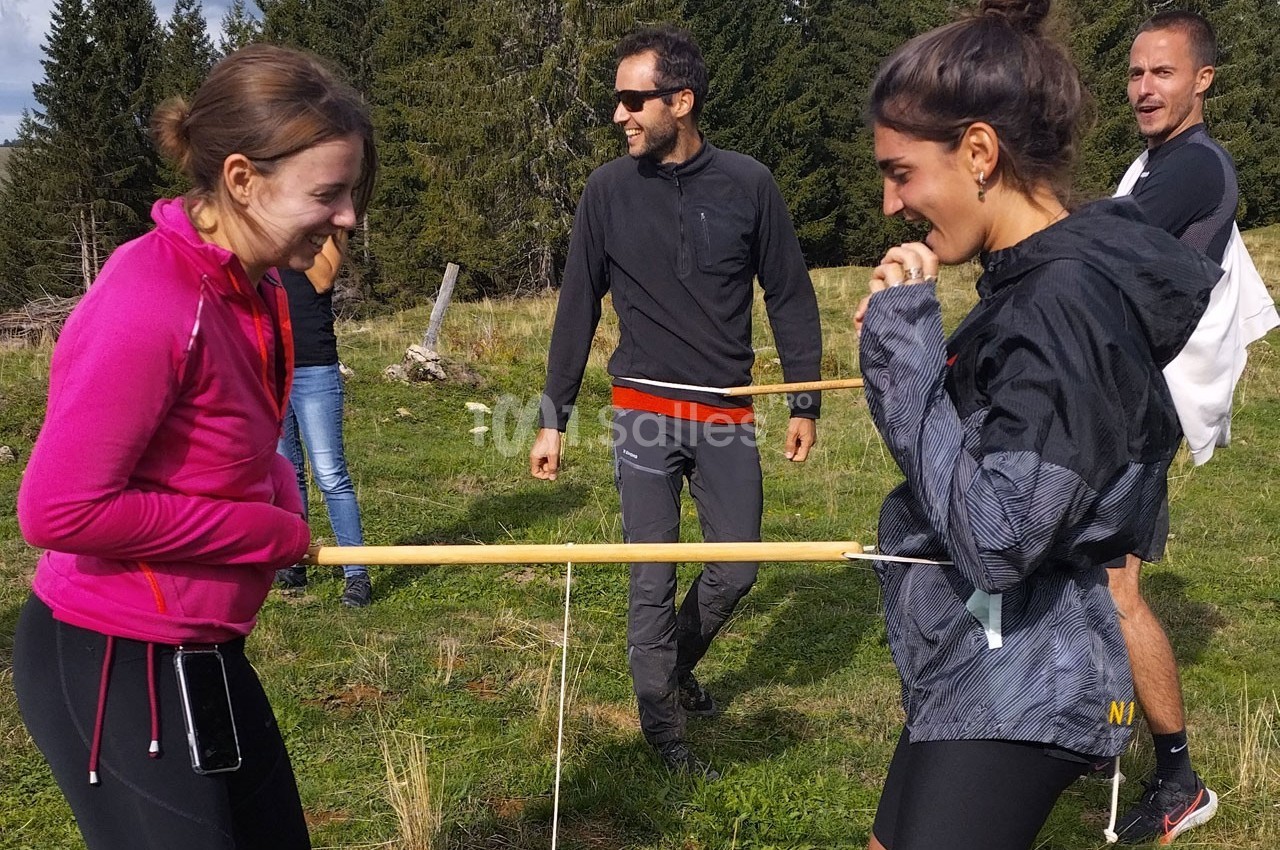 Deux femmes rient en participant à un jeu d'équilibre avec des bâtons, sous le regard de deux hommes en plein air.