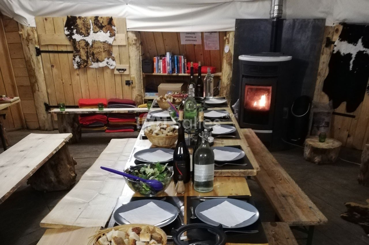 Table en bois dressée avec des assiettes, du pain et des boissons, dans une salle chaleureuse avec poêle à bois allumé.