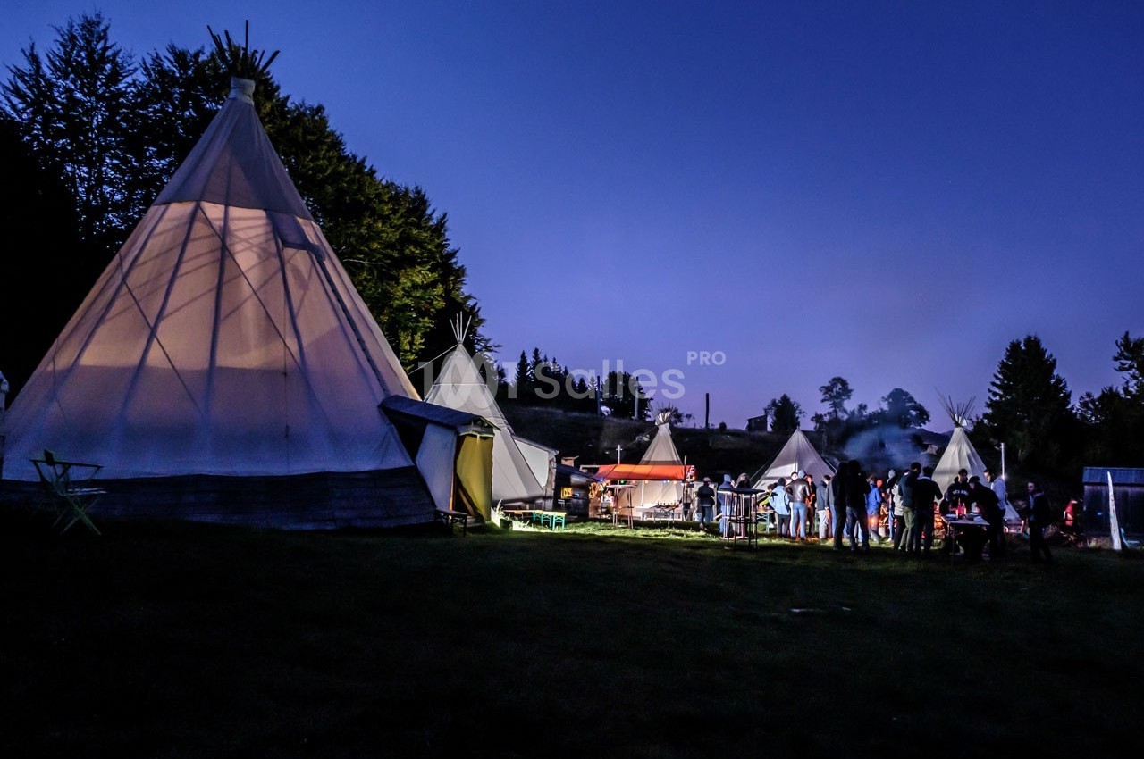Des tipis éclairés au crépuscule avec un groupe de personnes rassemblées autour d'une zone animée en plein air.