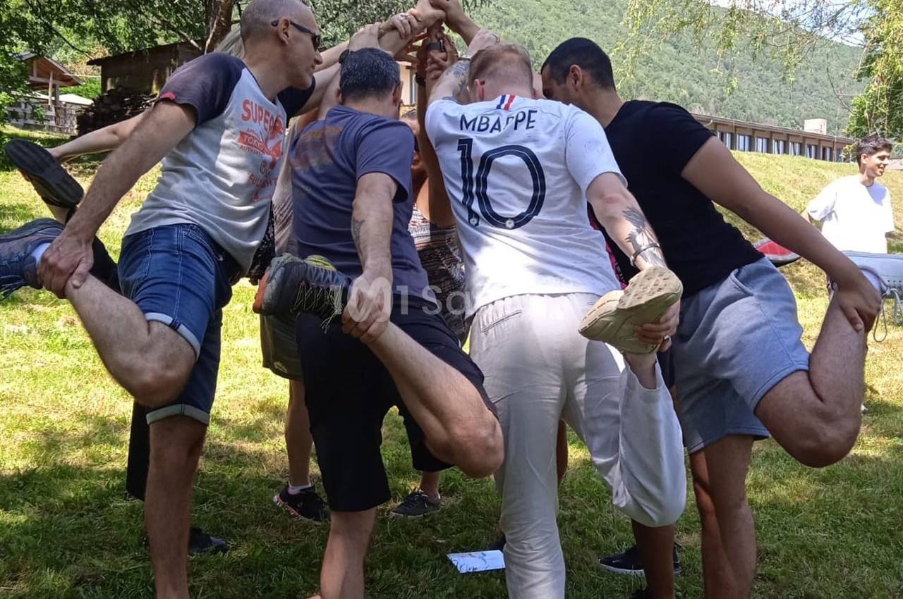 Un groupe de personnes en cercle, debout sur une jambe, se tenant par les bras dans un parc verdoyant.