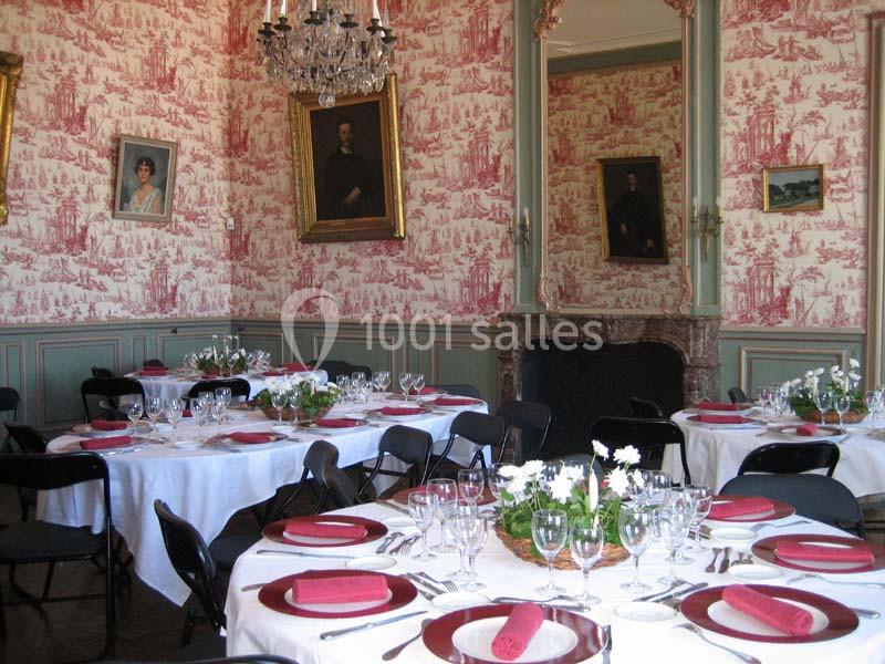 Salle élégante avec tables rondes dressées, nappes blanches, serviettes rouges, décor mural classique et cheminée ornée.