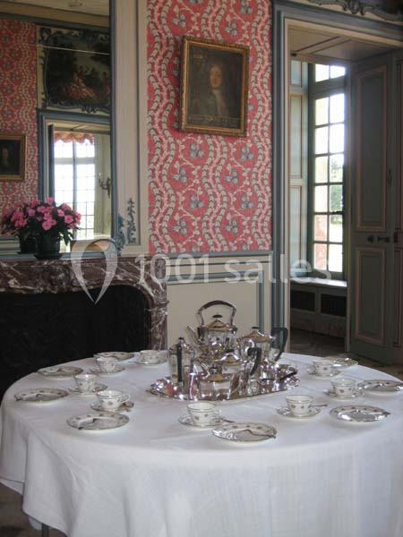 Table dressée avec un service à thé en porcelaine et en argent dans une pièce au décor classique avec cheminée et fenêtre.